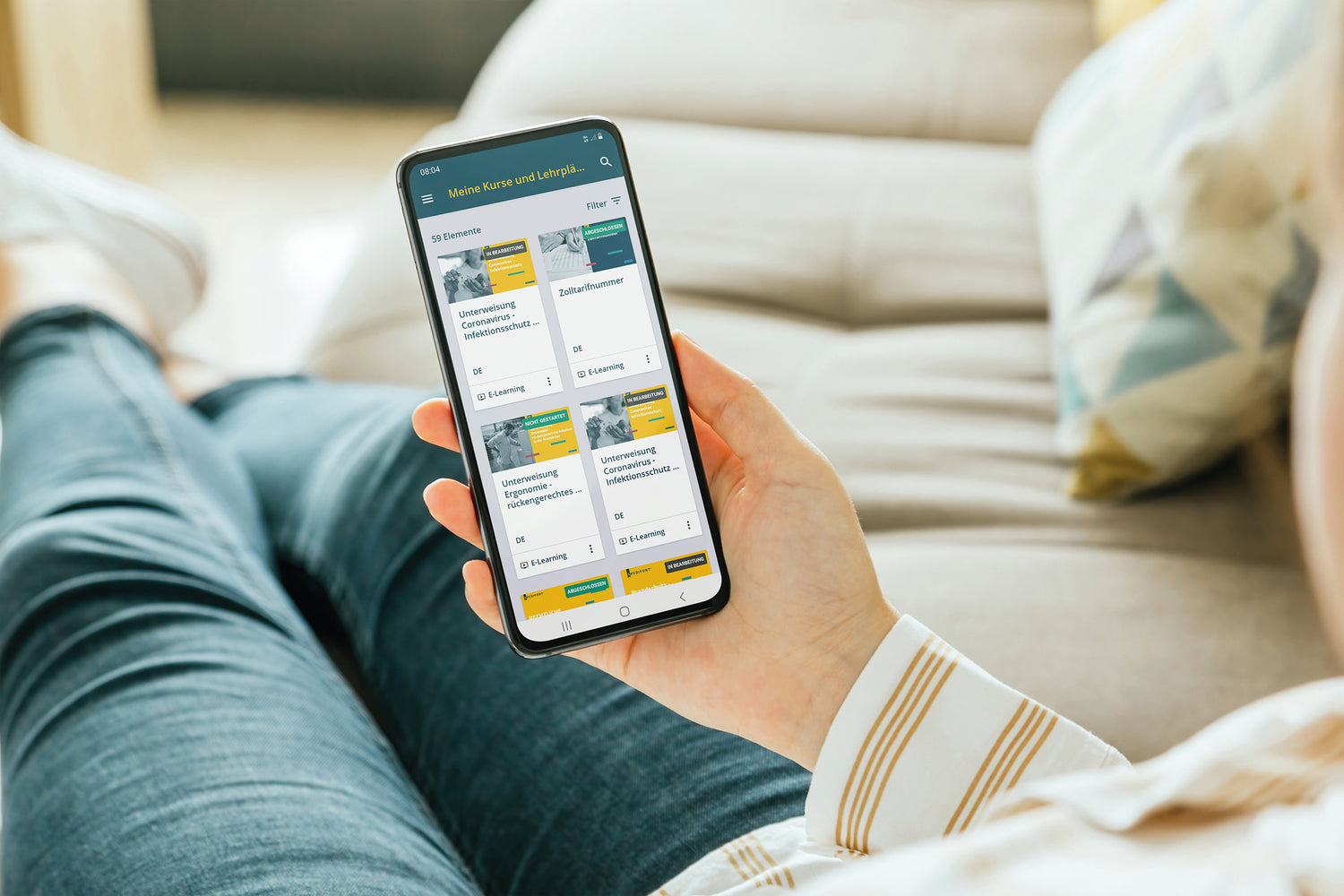 Person hält auf einem Sofa ein Smartphone in der Hand, auf dem die Spedifort-App mit einer Übersicht von E-Learning-Kursen angezeigt wird.